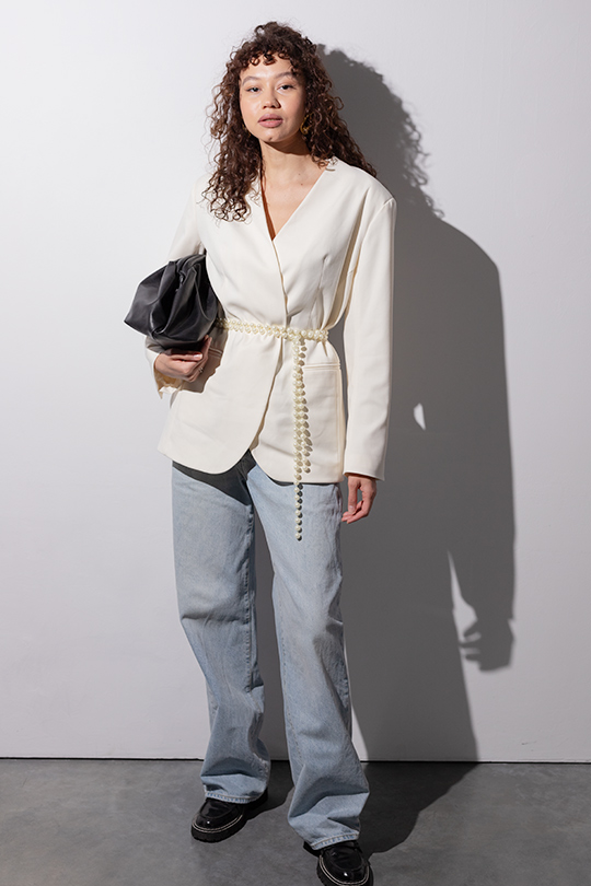 Девушка в белом жакете MUA с жемчужным поясом и светлыми джинсами держит в руке черную сумку-баул.